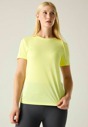 Mujer con largo cabello rubio que lleva una camiseta de manga corta de color amarillo brillante y leggings oscuros, sosteniendo el dobladillo de la camiseta con una mano.