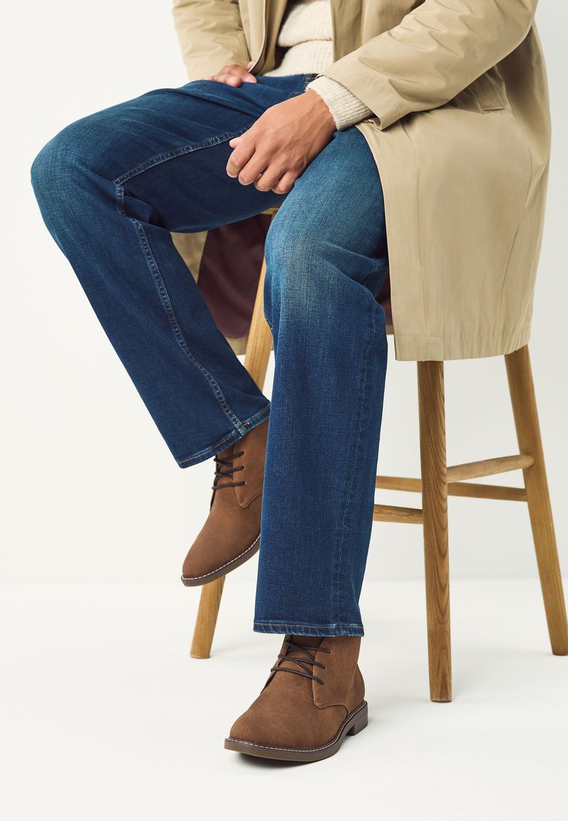 Person sitzt auf einem Holzschemel, trägt blaue Jeans, braune Schnürstiefel, einen beigen Trenchcoat und einen hellen Pullover.