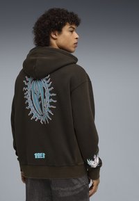 Νεαρός άνδρας με σκούφο φούτερ σε σκούρο χρώμα, με μπλε και ροζ σχεδιαστικά στοιχεία στην πλάτη και στο μανίκι, στέκεται μπροστά σε απλό γκρι φόντο.