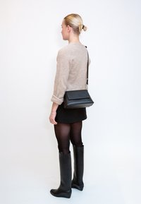 Γυναίκα με ξανθά μαλλιά σε κότσο, φοράει μπεζ πουλόβερ, μαύρη φούστα, μαύρες καλσόν και μαύρες μπότες έως το γόνατο, κρατώντας μια μαύρη τσάντα χιαστί.