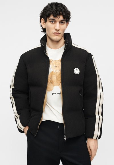 Νεαρός άνδρας με σγουρά μαλλιά που φοράει μαύρο μπουφάν με λευκές ρίγες και λευκό πουκάμισο με σχέδιο αρκουδάκι.