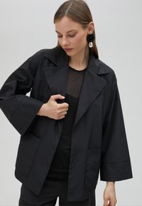 Černá lehká bunda s širokými rukávy, ostrým límcem a dvěma předními kapsami. Nosená přes upnutý černý síťovaný top.