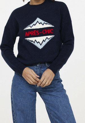 Persona con suéter azul marino con diseño de montaña y texto rojo "APRÈS-CHIC", combinado con jeans azules de talle alto, con las manos entrelazadas a la altura de la cintura.