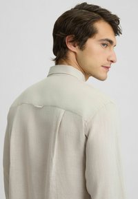 Camicia a maniche lunghe color beige chiaro, con colletto e pieghe sul retro, realizzata in un tessuto testurizzato che appare morbido e traspirante.