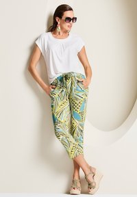 Weißes kurzärmliges Oberteil und grüne gemusterte Hose mit Blättern und geometrischen Formen. Plateausandalen mit strukturierten Riemen, übergroße Sonnenbrille.