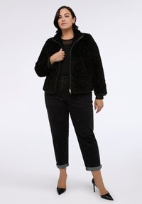 Donna in giacca di pelliccia sintetica nera, top nero, jeans a vita alta con delicata ricamata floreale, e tacchi a spillo neri, in piedi con sicurezza.