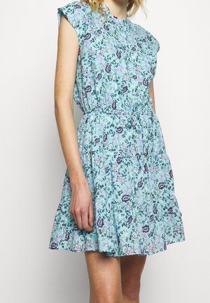 Frau trägt ein ärmelloses hellblaues Kleid mit floralem Muster und tailliert, steht vor einem einfarbigen Hintergrund.