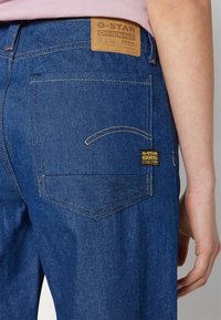 Niebieskie dżinsy z denimu z brązową skórzaną metką na tylnej części pasa oraz obszytymi tylnymi kieszeniami o zaokrąglonym kształcie.