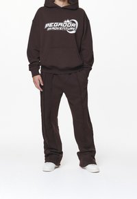 Bruine hoodie met een grafisch logo, gecombineerd met bijpassende bruine broek met zijstikseldetails. Witte sneakers maken de look compleet.