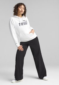 Felpa bianca con logo PUMA nero, dotata di tasca anteriore, abbinata a pantaloni larghi neri e scarpe bianche. Tessuto liscio, design casual.
