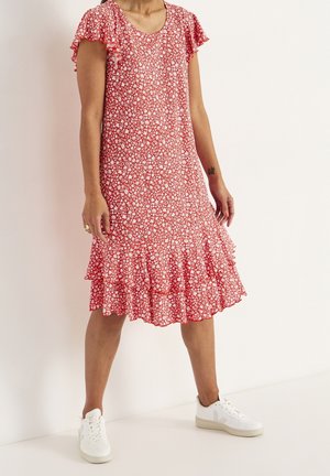 Donna che indossa un vestito rosso con stampa floreale fino al ginocchio, con maniche e orlo arricciati, abbinato a scarpe da ginnastica bianche, in piedi contro una parete semplice.