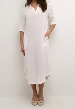 Kvinde iført en hvid midi-kjole med trekvartærmer, sidelommer og beige spidse flade sko, stående foran en ensfarvet baggrund.