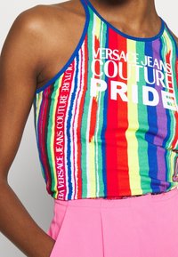 Personne portant un débardeur rayé coloré avec le texte "Versace Jeans Couture Pride" et un pantalon rose vif, main dans la poche.