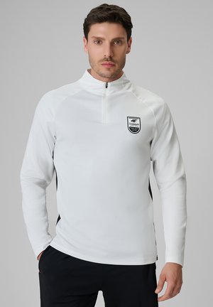 Mężczyzna noszący biały sportowy top z długim rękawem i zamkiem błyskawicznym, z czarnym logo na lewej piersi i czarne spodnie.