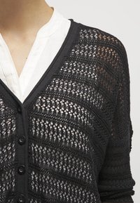 Cárdigan negro de punto con un patrón de tejido abierto, escote en forma de V y cierre de botones. Cuenta con mangas largas y un acabado texturizado.