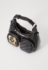 Černá kožená kabelka se dvěma předními kapsami na zip, zlatým kováním, značkovou emblémou a krátkým uchem na jednobarevném pozadí.