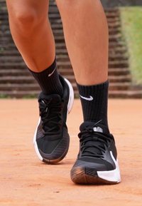 Black athletic shoes with a textured fabric upper and white accents, paired with black ribbed socks. Focus on footwear in motion on a court surface.