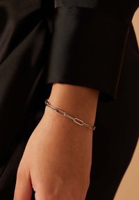 Zilveren armband met verlengde schakels, met een gladde oppervlakte. Gedragen om een pols tegen een zwart stoffen achtergrond.