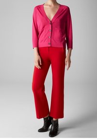 Roze cardigan met V-hals, driekwart mouwen en twee voorzakken, gecombineerd met rode brede broek en zwarte enkellaarsjes.