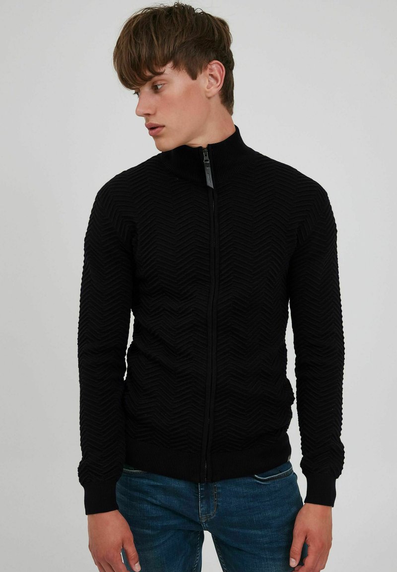 Sudadera negra con cremallera y un patrón en zigzag texturizado, cuello alto y puños de canalé, combinada con jeans de mezclilla azul.