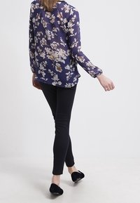 Blouse en shet de couleur navy à motif floral, avec des manches longues, associée à un pantalon noir slim-fit et des chaussures plates noires. Vue de dos affichée.