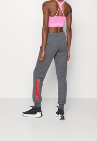 Grå joggingbukser med elastisk taljebånd, der har en stor rød "adidas"-logo på venstre ben. Matcher med en lyserød atletisk crop top og sorte sko.