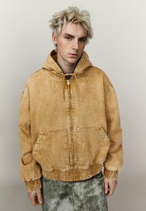 Jeune personne aux cheveux blonds courts et en pointe, portant un sweat à capuche marron délavé et surdimensionné avec fermeture éclair, et un pantalon vert tie-dye, se tenant devant un fond uni.