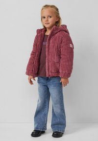 Plüschige, gerippte, burgunderrote Pufferjacke mit Kapuze, kombiniert mit einem dunklen T-Shirt und hellblauen Schlagjeans; schwarze Schuhe.