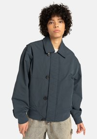 Young person with curly hair wearing a dark blue buttoned jacket and light blue jeans, standing against a plain white background.