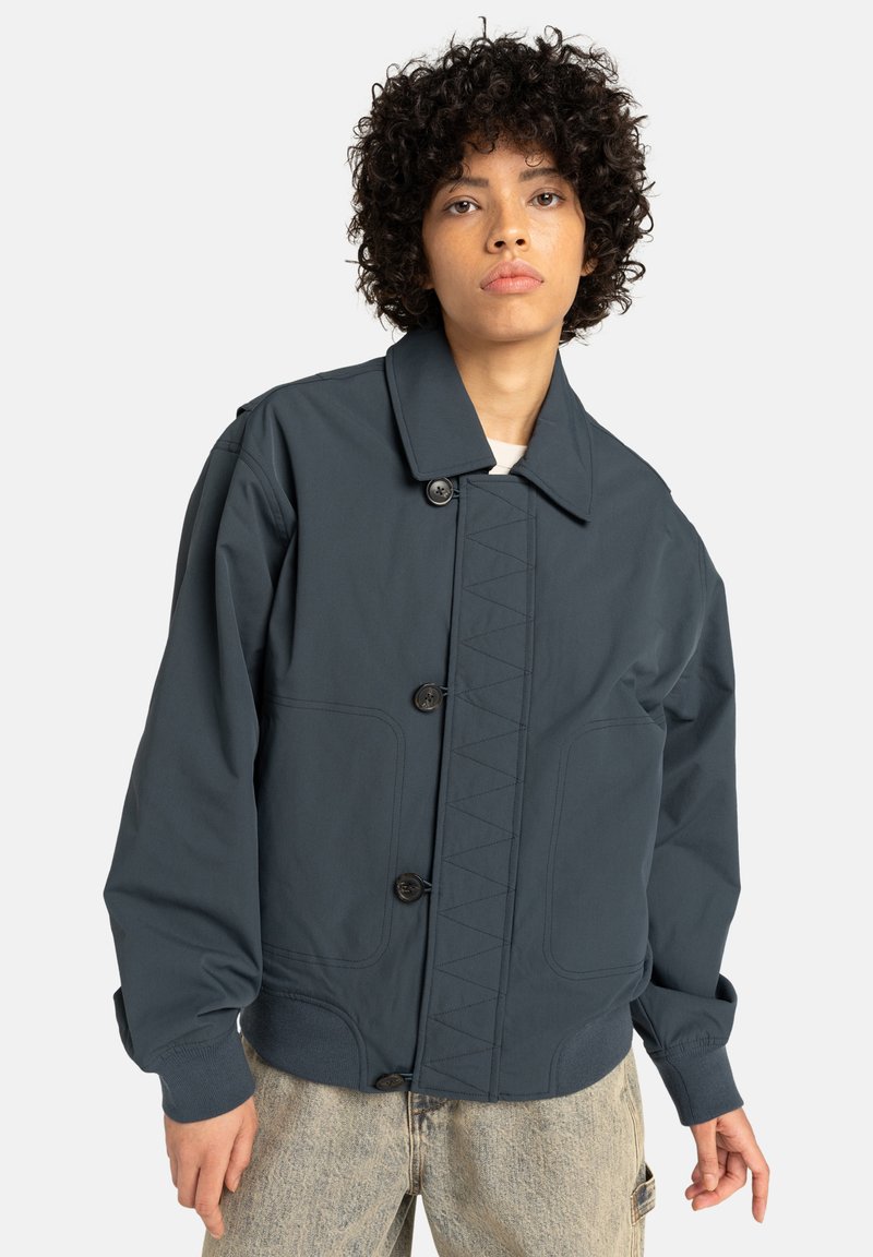 Young person with curly hair wearing a dark blue buttoned jacket and light blue jeans, standing against a plain white background.