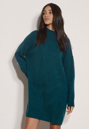Giovane donna con lunghi capelli scuri che indossa un abito maglione largo color teal, in piedi con una mano sul fianco su uno sfondo chiaro uniforme.