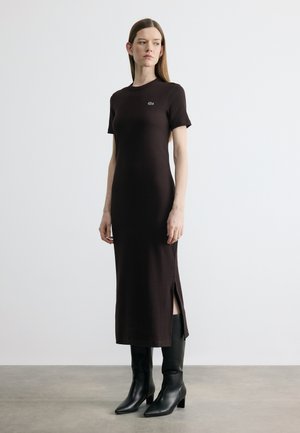 Vrouw draagt een zwarte midi-jurk met korte mouwen en een split aan de zijkant, gecombineerd met zwarte laarzen met hak, staand tegen een effen lichte achtergrond.