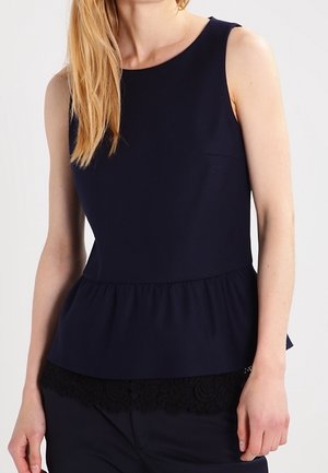 Vrouw draagt een mouwloze navy peplum top met kanten rand detail aan de zoom, gecombineerd met een bijpassende navy broek.