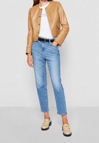 Kvinna klädd i en beige läderjacka, vit skjorta, ljusblå jeans, svart bälte och beige loafers som står med en hand i fickan.