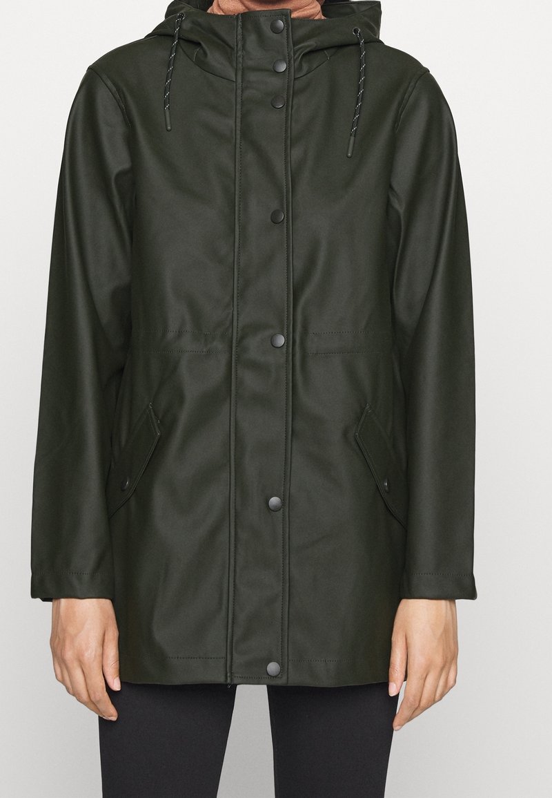 Veste imperméable vert foncé avec capuche, boutons-pression à l'avant, poches à rabat inclinées et cordons, portée sur un pantalon noir.