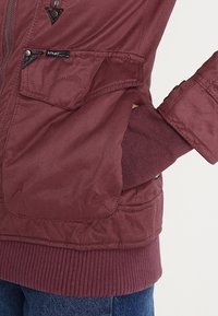 Poche de veste bordeaux avec bouton-pression et petite étiquette, main partiellement à l'intérieur, poignets et ourlet côtelés, associée à un jean en denim bleu.