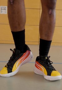 Sorte og gule sportsko med gradient røde og orange detaljer, med hvide striber og strukturerede gummisåler, båret med sorte sokker.