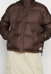 Braune Daunenjacke mit einem Frontreißverschluss, gerippten Bündchen und seitlichen Taschen; verfügt über ein Logo-Patch in der Nähe des Saums. Das Material wirkt glatt und gepolstert.