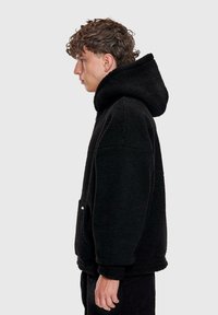 Giovane uomo con capelli ricci che indossa una felpa con cappuccio nera e texture, in profilo laterale su uno sfondo chiaro e semplice.