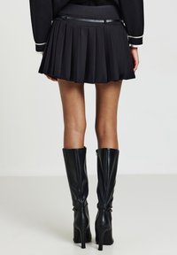 Femme portant une mini-jupe plissée noire avec une fine ceinture, associée à des bottes en cuir noirs à talons hauts jusqu'aux genoux, vue de dos.