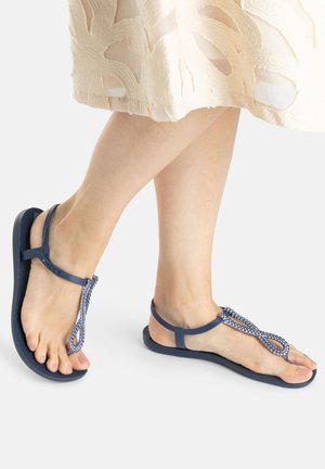 Blue sandals with a flat sole, featuring a braided upper design and a T-strap style. Soft texture, minimalistic hardware accents visible.