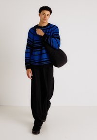 Pull rayé bleu et noir avec un col rond et une coupe ample, associé à un pantalon noir ample et un sac à bandoulière noir.