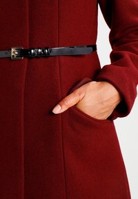 Mano nella tasca di un cappotto di lana bordeaux con una cintura nera slim che presenta una fibbia dorata in vita.