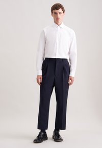 Biała koszula na guziki z kieszenią na piersi, zestawiona z granatowymi spodniami z wysokim stanem. Modelka ma na sobie czarne sznurowane buty. Gładkie tło.