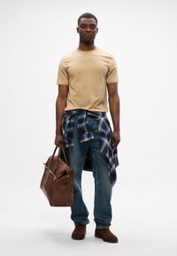 Camisa de manga corta color beige, camisa a cuadros azul atada a la cintura, jeans azules, botas de cuero marrón y un bolso de cuero marrón.