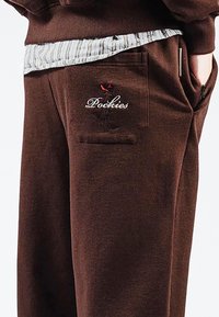 Braune Jogginghosen mit einer Gesäßtasche, die ein gesticktes Logo "Pochies" und ein Figurendesign zeigt. Der Stoff wirkt weich und strukturiert.