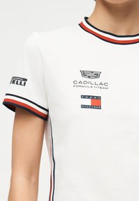 Λευκό μπλουζάκι με κόκκινο, άσπρο και μπλε ριγέ γιακά και άκρες μανικιών, με λογότυπα της ομάδας Formula 1 της Cadillac και της Tommy Hilfiger.