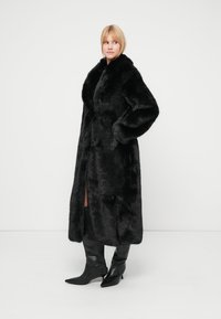 Svart kåpe i kunstpels med bred krage og lange ermer, med sidespalter og en glatt tekstur, brukt sammen med tettsittende svarte støvler som rekker opp til knærne.