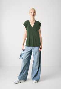 Grön V-ringad topp med korta ärmar, kombinerad med vida ljusblå jeans och vita sneakers. Håller en ljusblå handväska.