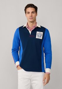 Man wearing a blue and navy long-sleeve rugby shirt with a white collar, patch logo, and white pants, standing against a plain background.